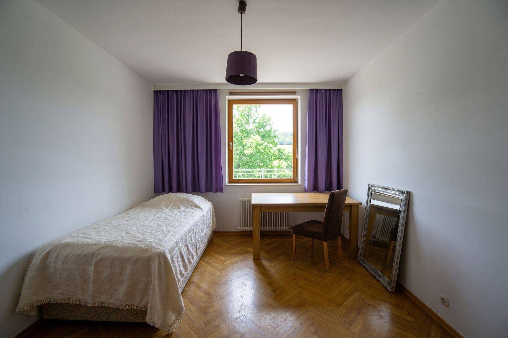 Einfaches Schlafzimmer mit Einzelbett, Holzboden und Fenster mit lila Vorhängen.