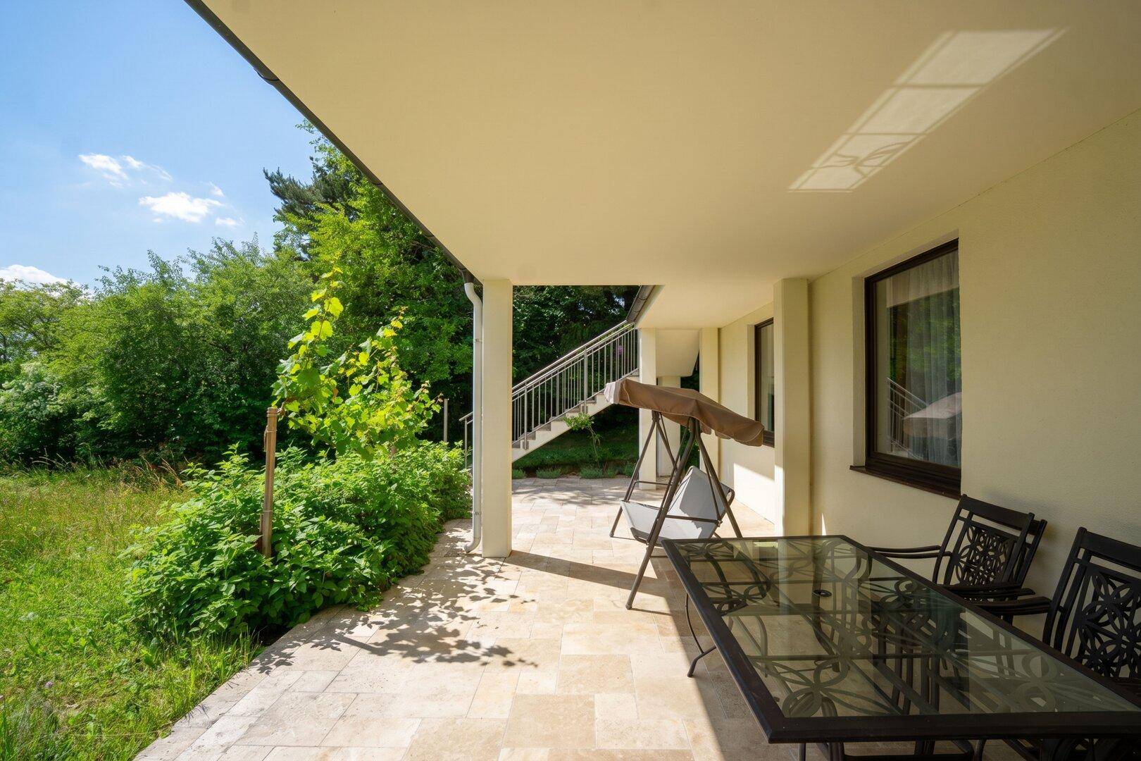 Überdachte Terrasse mit Esstisch und Stühlen, umgeben von üppigem Grün.