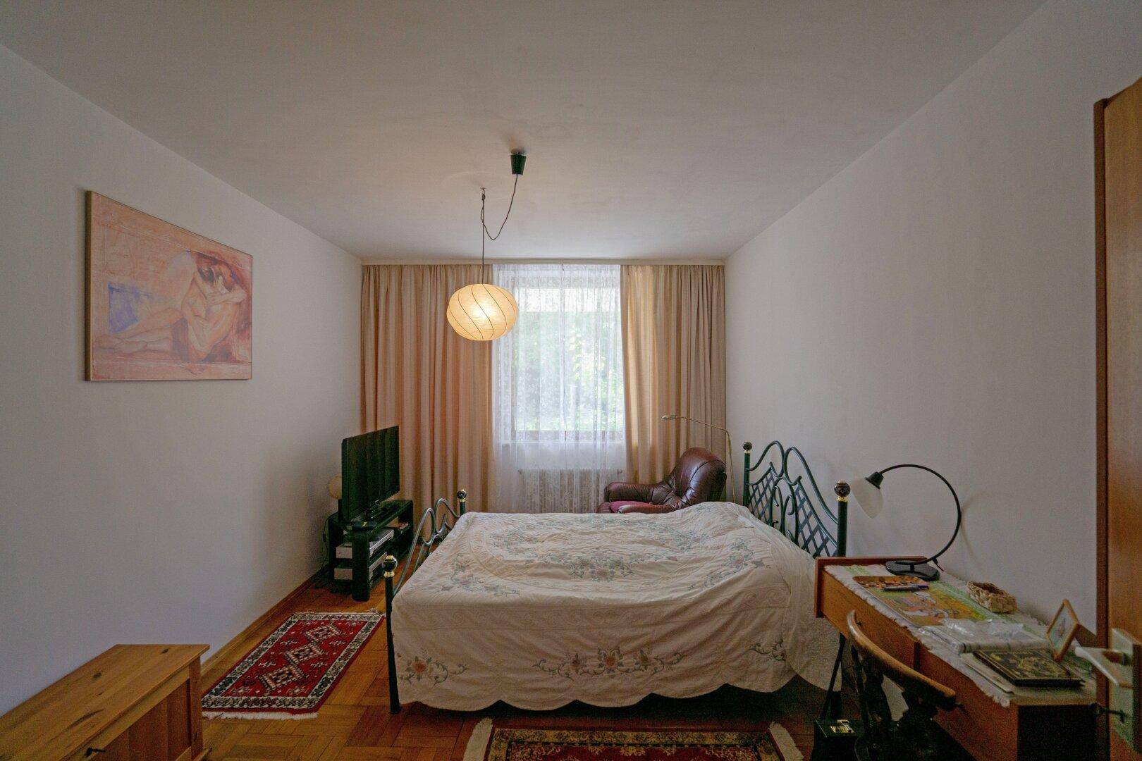 Schlafzimmer mit Metallbettgestell, Holzboden und Fenster mit Vorhängen.
