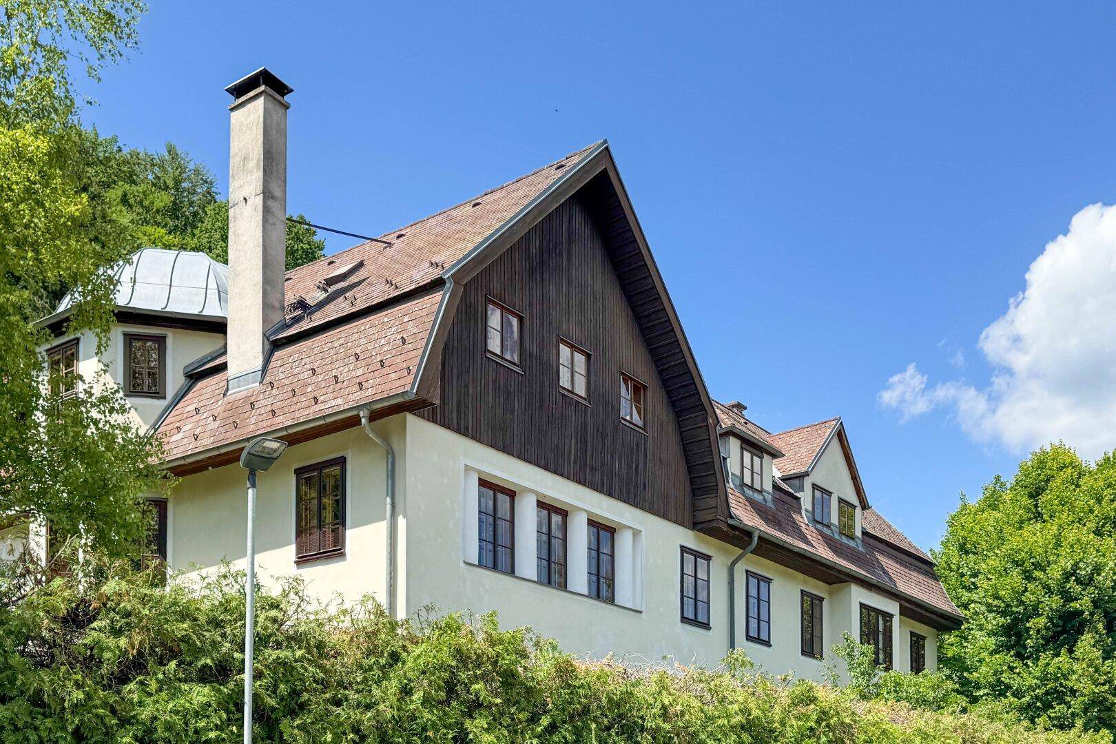 Traditionelles Haus mit markantem Dach, weißer Fassade und Holzelementen, umgeben von viel Grün.