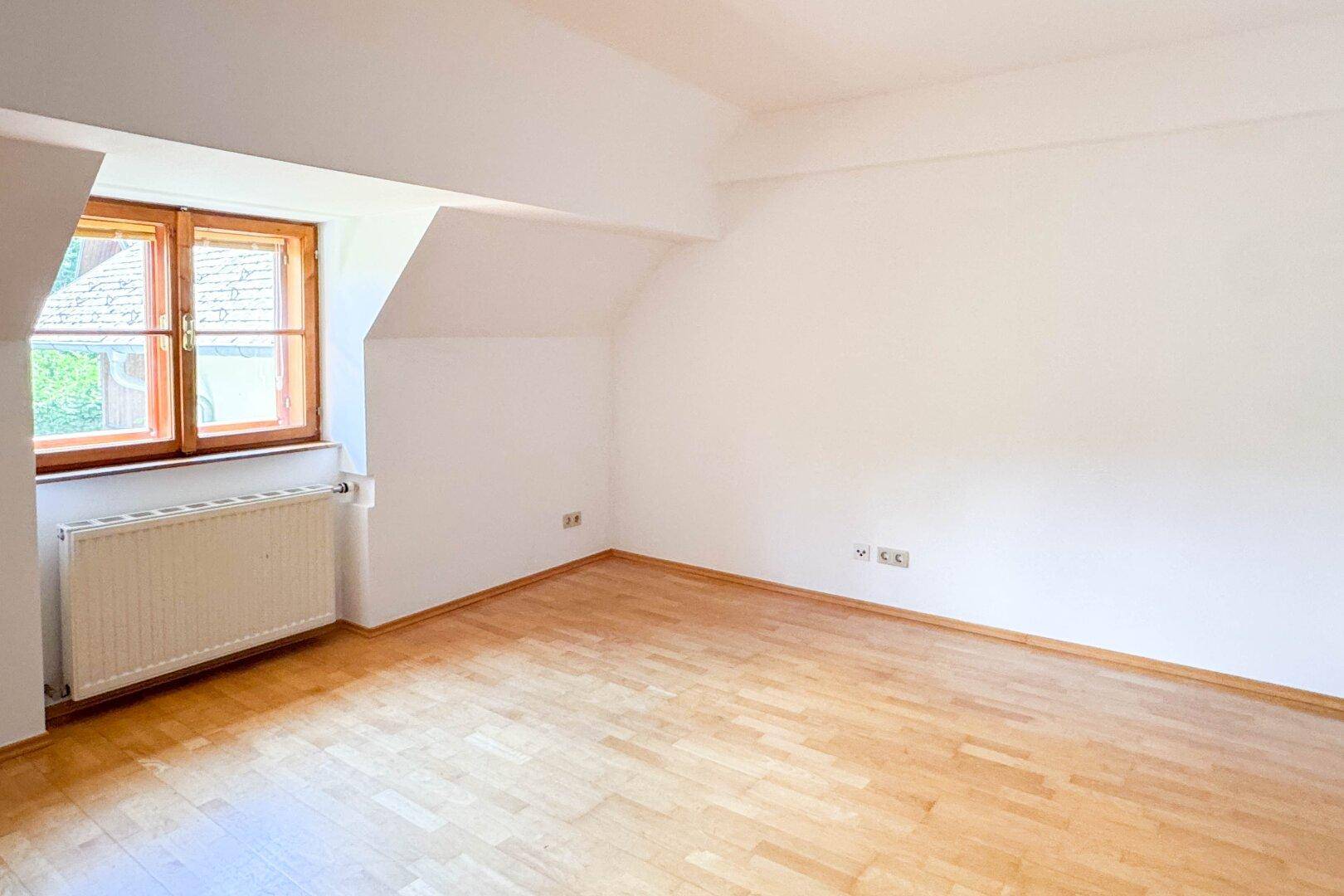 Ruhiges Zimmer mit Holzboden, weiß gestrichenen Wänden und einem Fenster im Dachgeschoss.