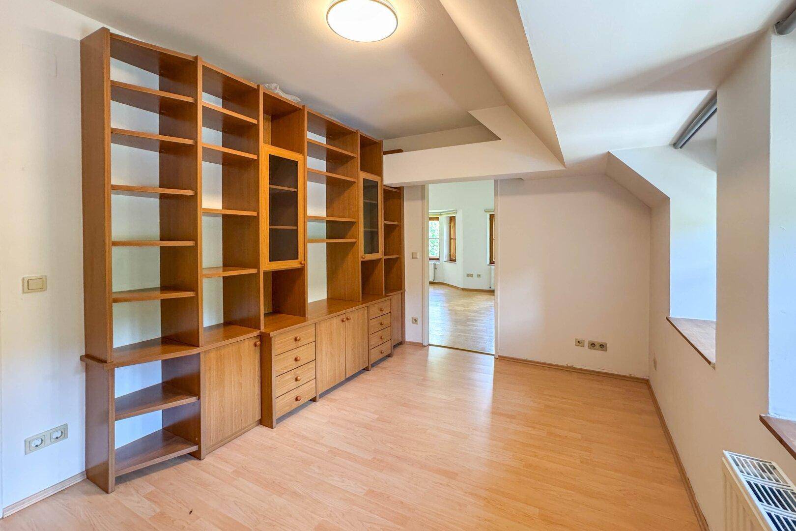 Zimmer mit großem Einbauregal aus Holz und Holzboden, mit Zugang zu einem weiteren Raum.