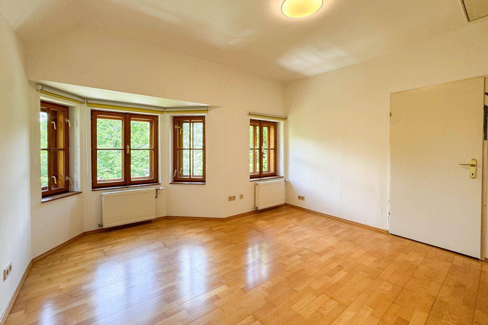 Helles Zimmer mit Holzboden und einem Erker mit mehreren Fenstern, die viel Licht hereinlassen.