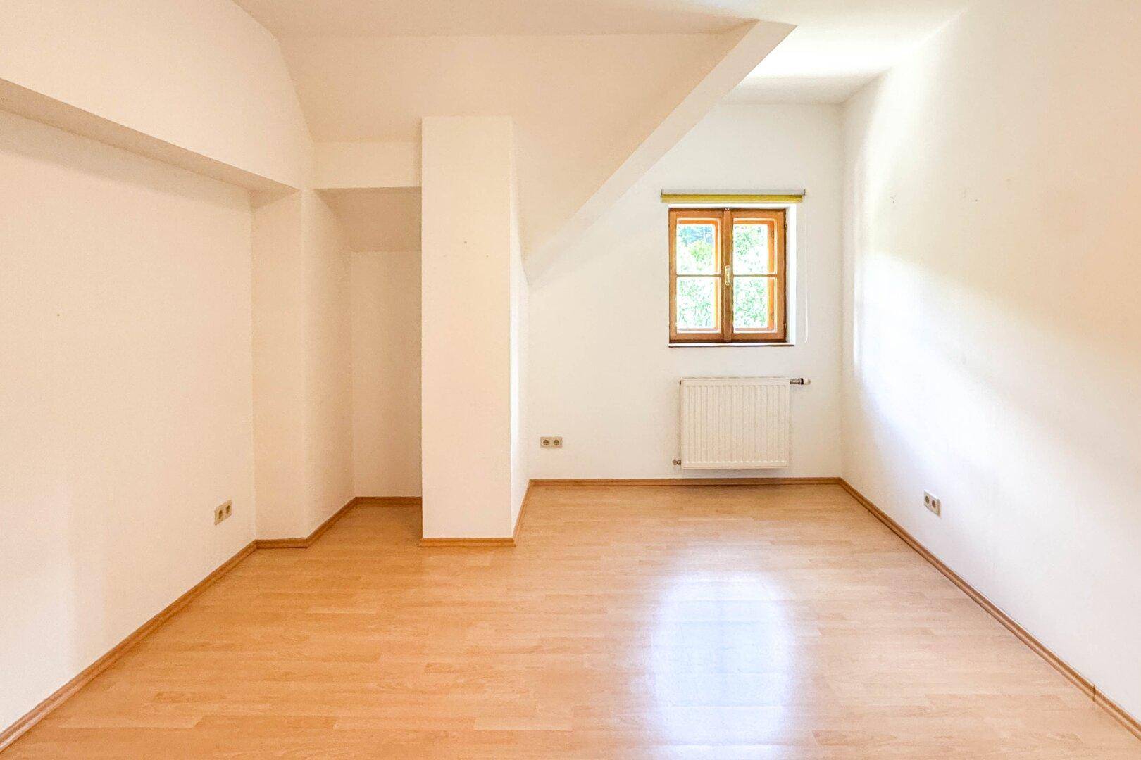 Helles Zimmer mit Holzboden, weiß gestrichenen Wänden und einem Fenster im Dachgeschoss.