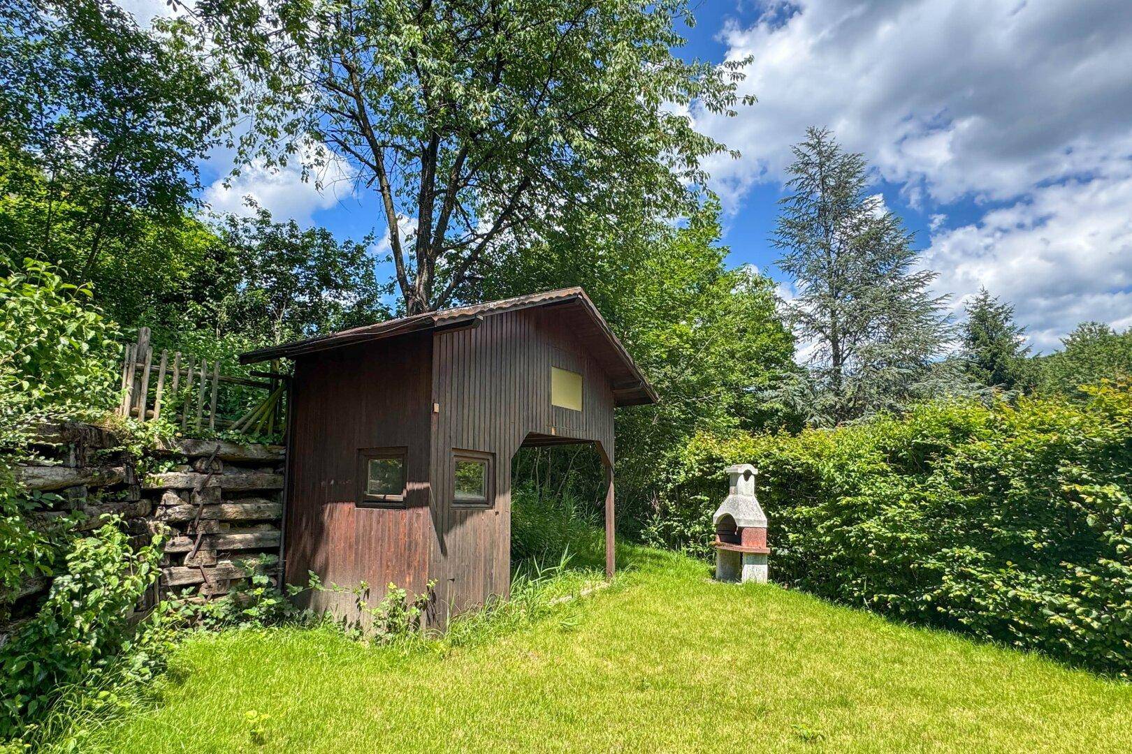 Idyllischer Gartenbereich mit einem Holzschuppen, Grillplatz und viel Grünfläche unter Bäumen.