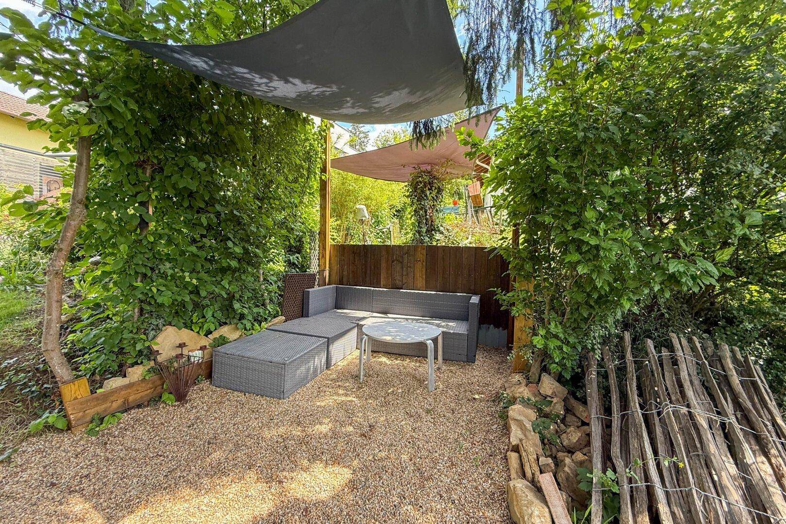Gemütlicher, schattiger Sitzbereich im Garten mit Lounge-Möbeln und Kiesboden.