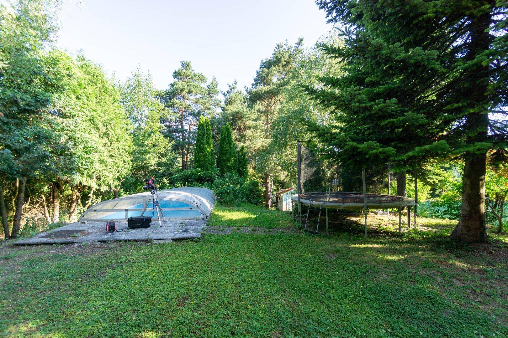 Weitläufiger Gartenbereich mit überdachtem Swimmingpool und großem Trampolin, umgeben von Bäumen.