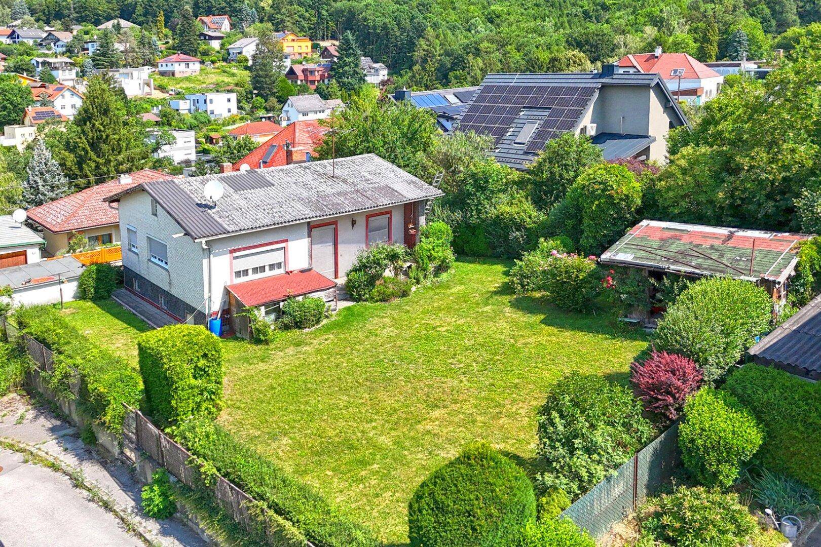 Luftaufnahme des Einfamilienhauses mit großem Garten und umliegender Wohnsiedlung in grüner Hügellandschaft.