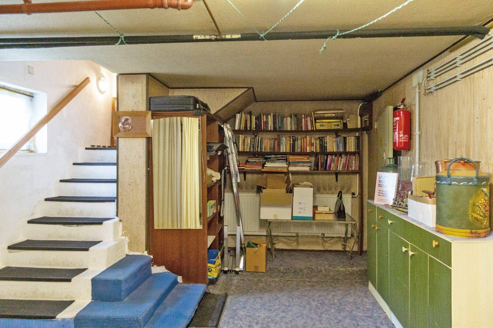 Kellerraum mit Treppenzugang, Bücherregalen und sichtbaren Rohren an der Decke.