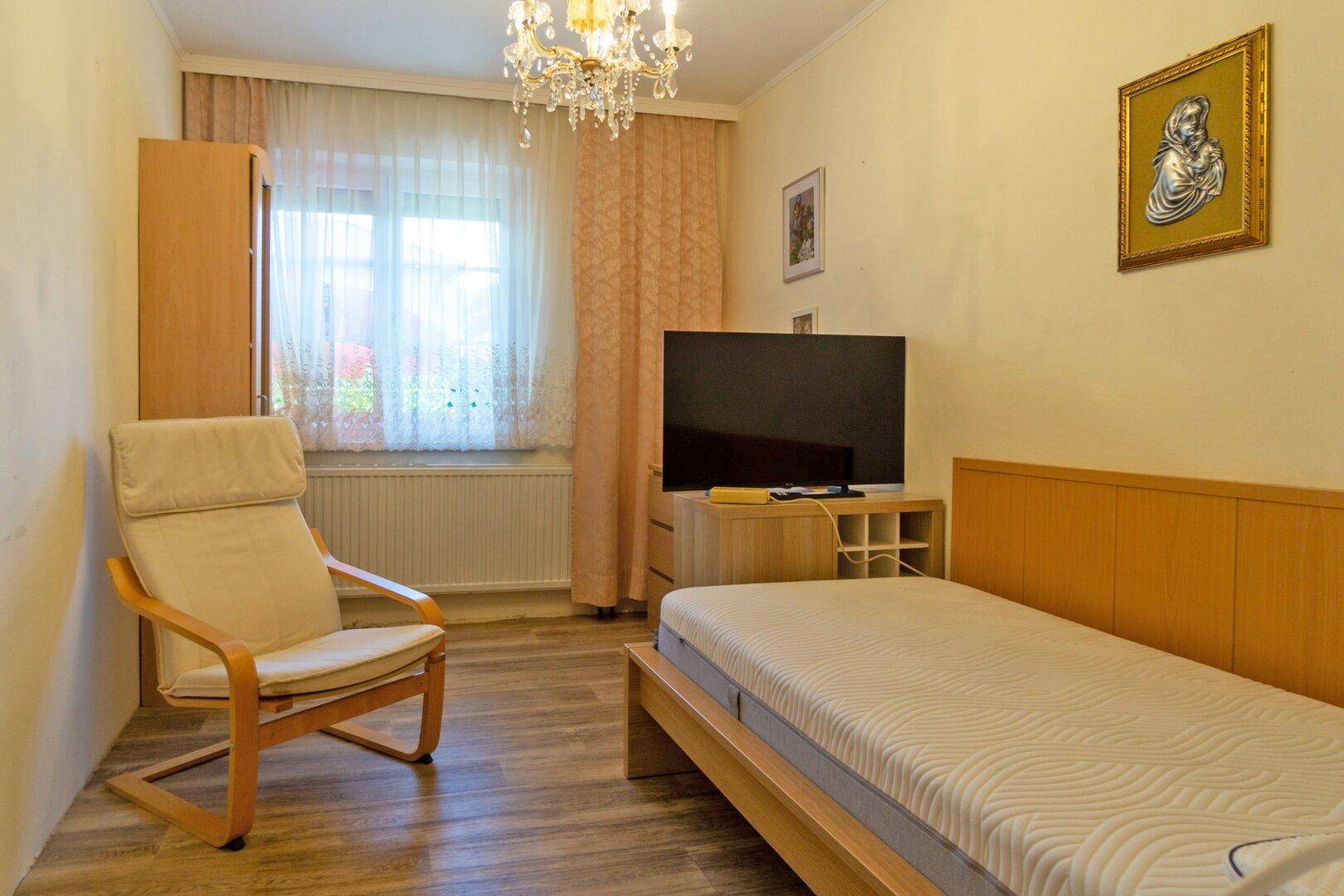 Helles Schlafzimmer mit Einzelbett, Sessel und Fernseher, dekoriert mit einem Kronleuchter.