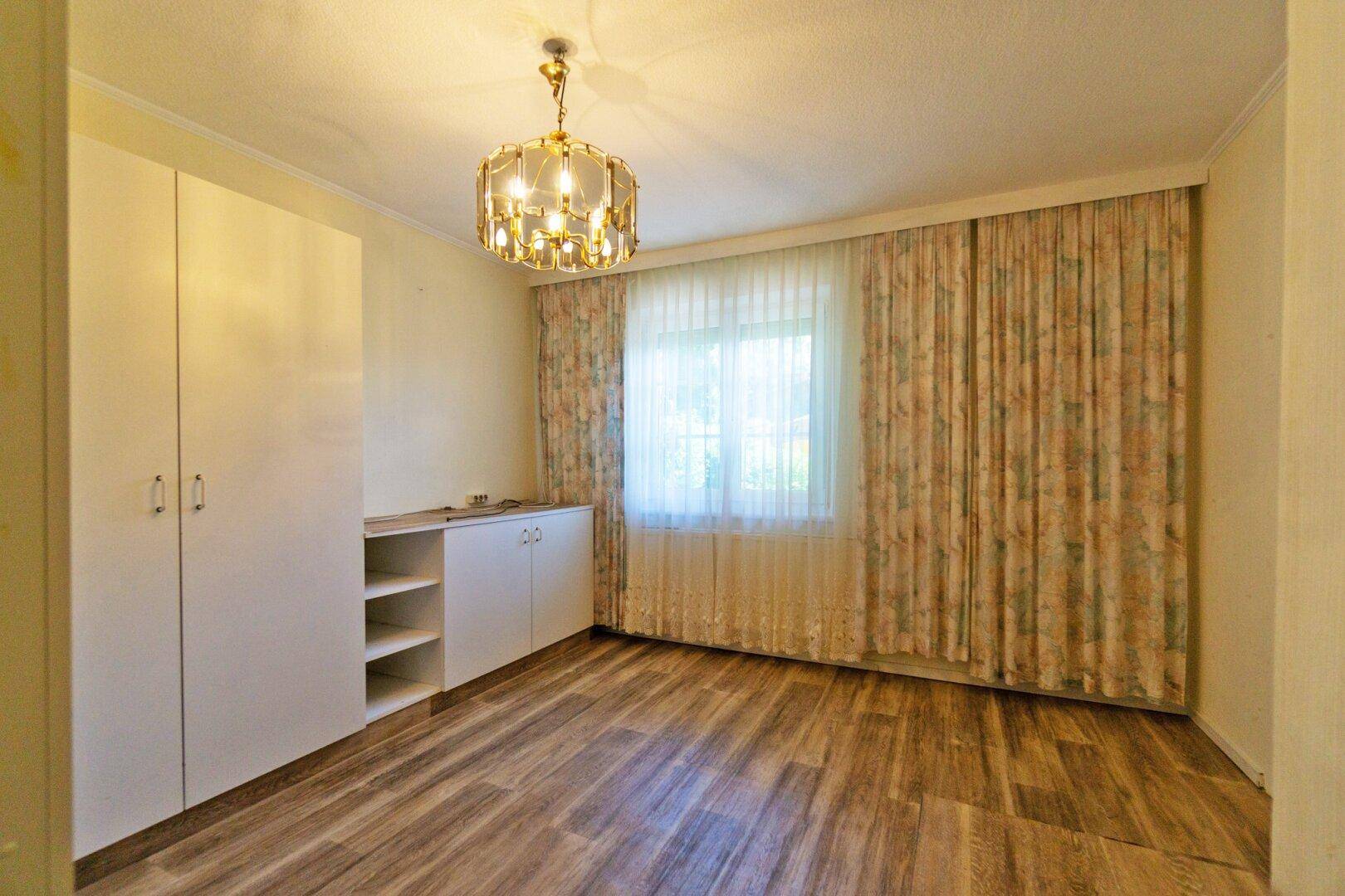 Schlafzimmer mit Laminatboden, weißen Schränken und einem großen Fenster mit Vorhängen.