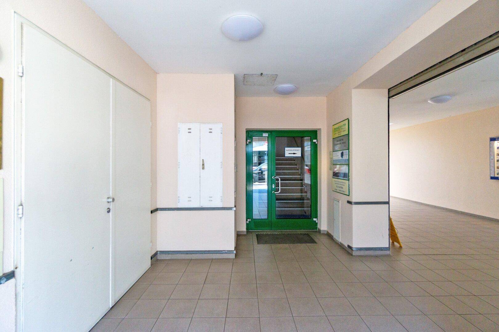 Heller Eingangsbereich mit Fliesenboden, weißen Wänden und einer grünen Tür, die zum Treppenhaus führt.