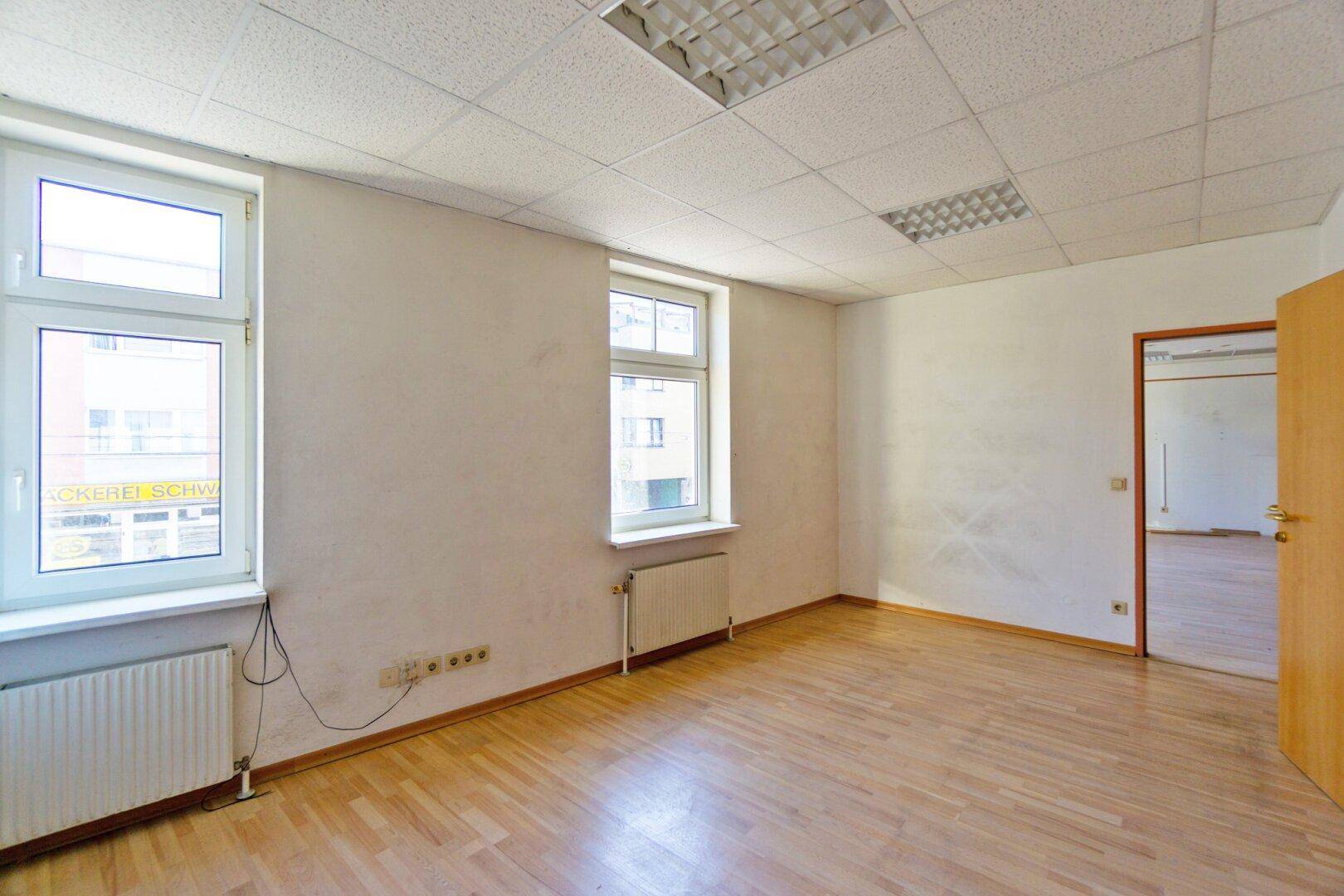 Heller Büroraum mit Holzfußboden, großen Fenstern und einer Tür, die zu einem weiteren Bereich führt.