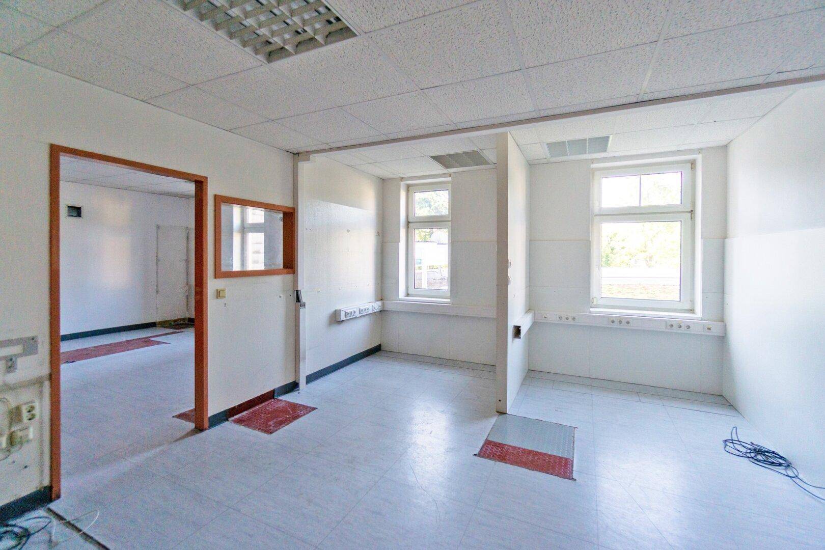Leerer Raum mit hellem Fliesenboden, Fenstern und einer Tür, geeignet als Büro oder Behandlungszimmer.