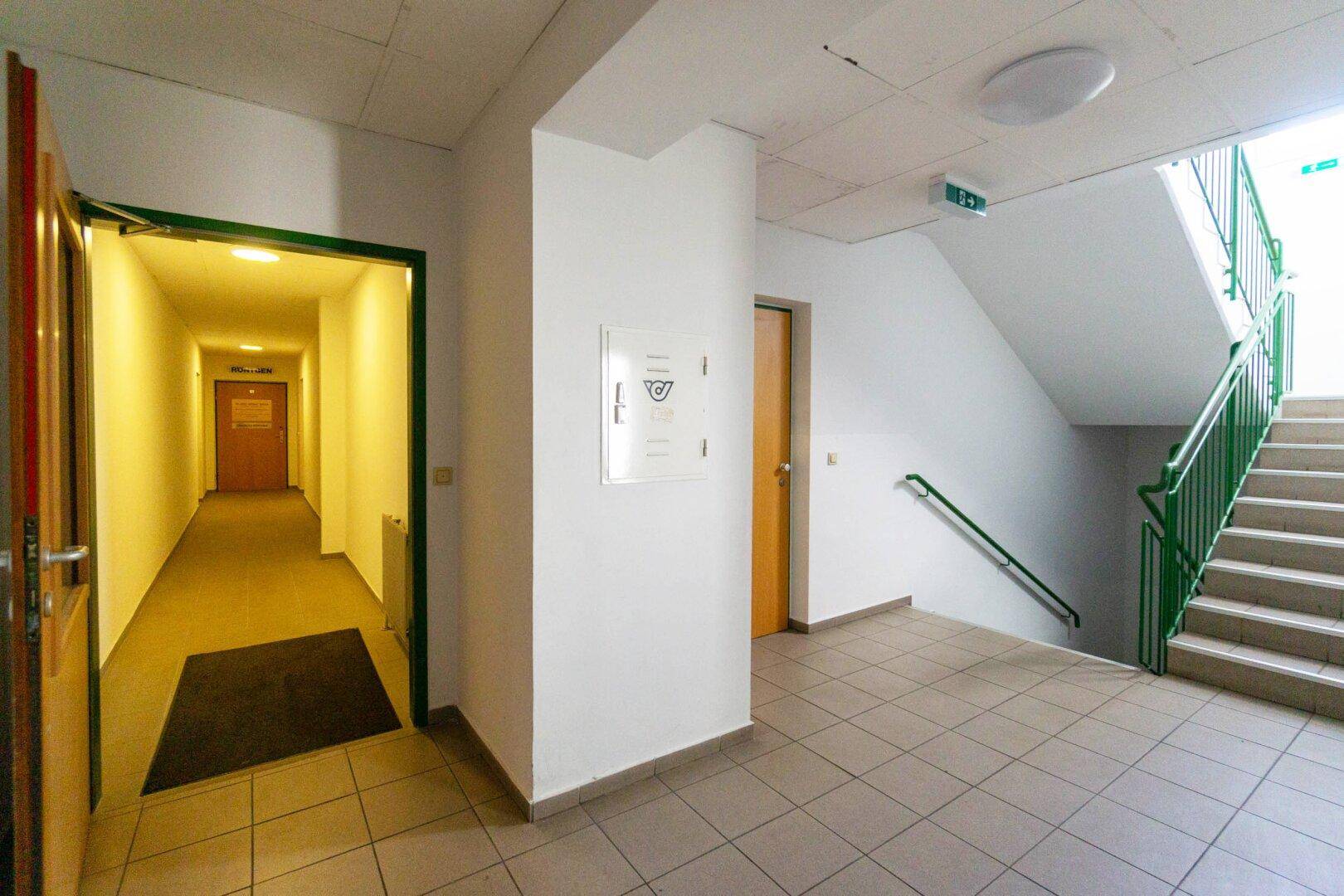 Langer Flur mit Fliesenboden, der zu einem Treppenhaus mit grünen Geländern und weiteren Türen führt.