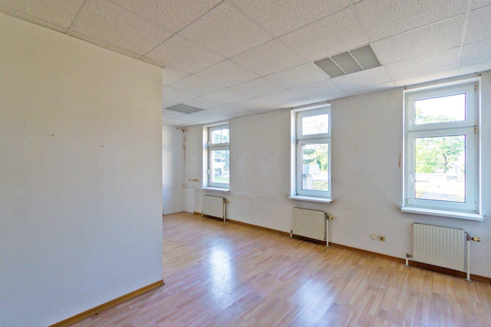 Geräumiger, leerer Büroraum mit Holzboden, drei großen Fenstern und Heizkörpern.