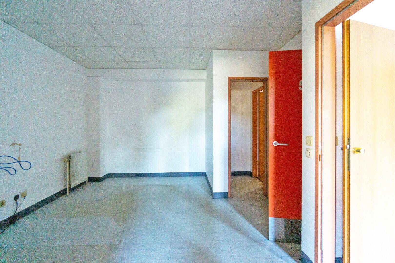 Leerer Raum mit Fliesenboden, weißen Wänden und einer auffälligen roten Tür, die zu weiteren Räumen führt.