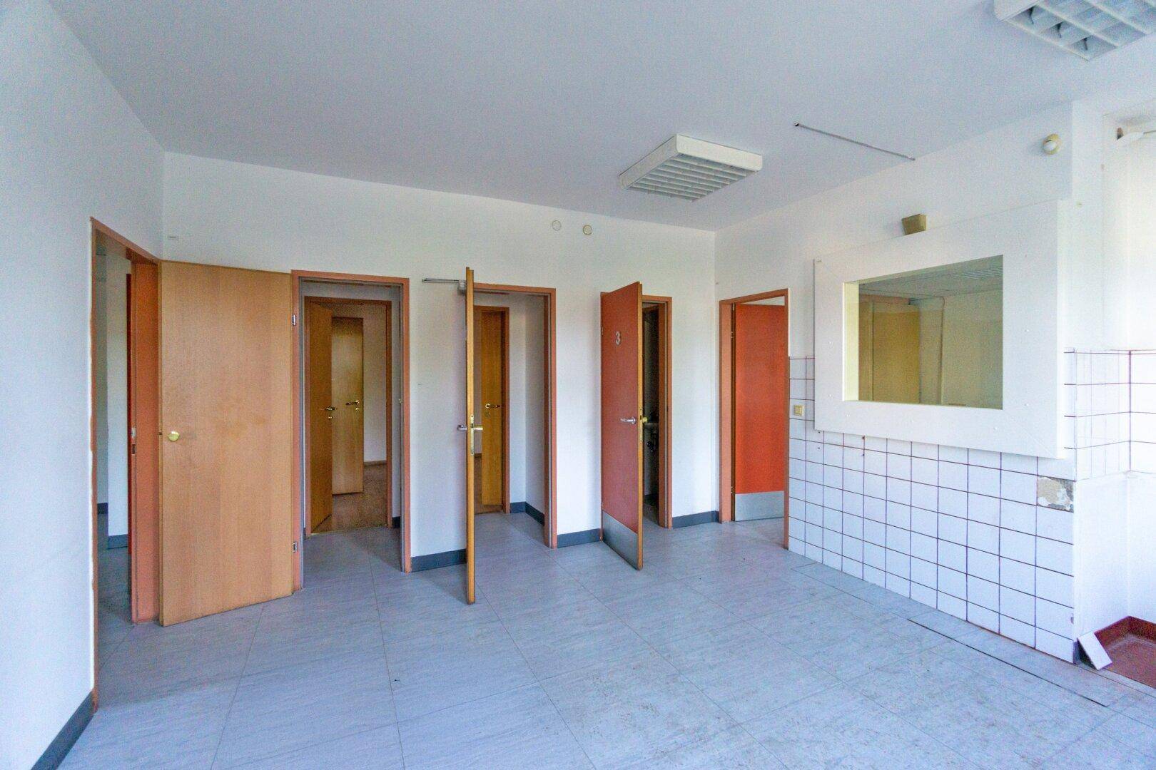 Geräumiger Raum mit Fliesenboden und mehreren Holztüren, der als Wartebereich oder Büro genutzt werden könnte.