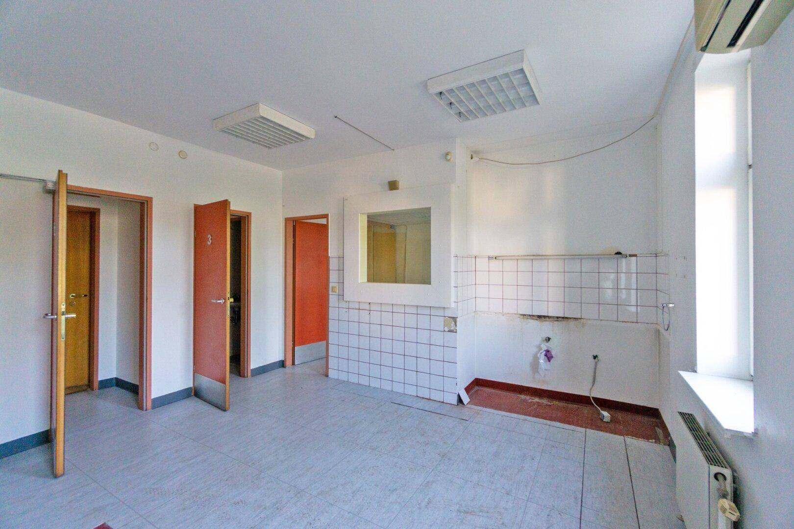 Geräumiger Raum mit Fliesenboden und mehreren Holztüren, der als Wartebereich oder Büro genutzt werden könnte.