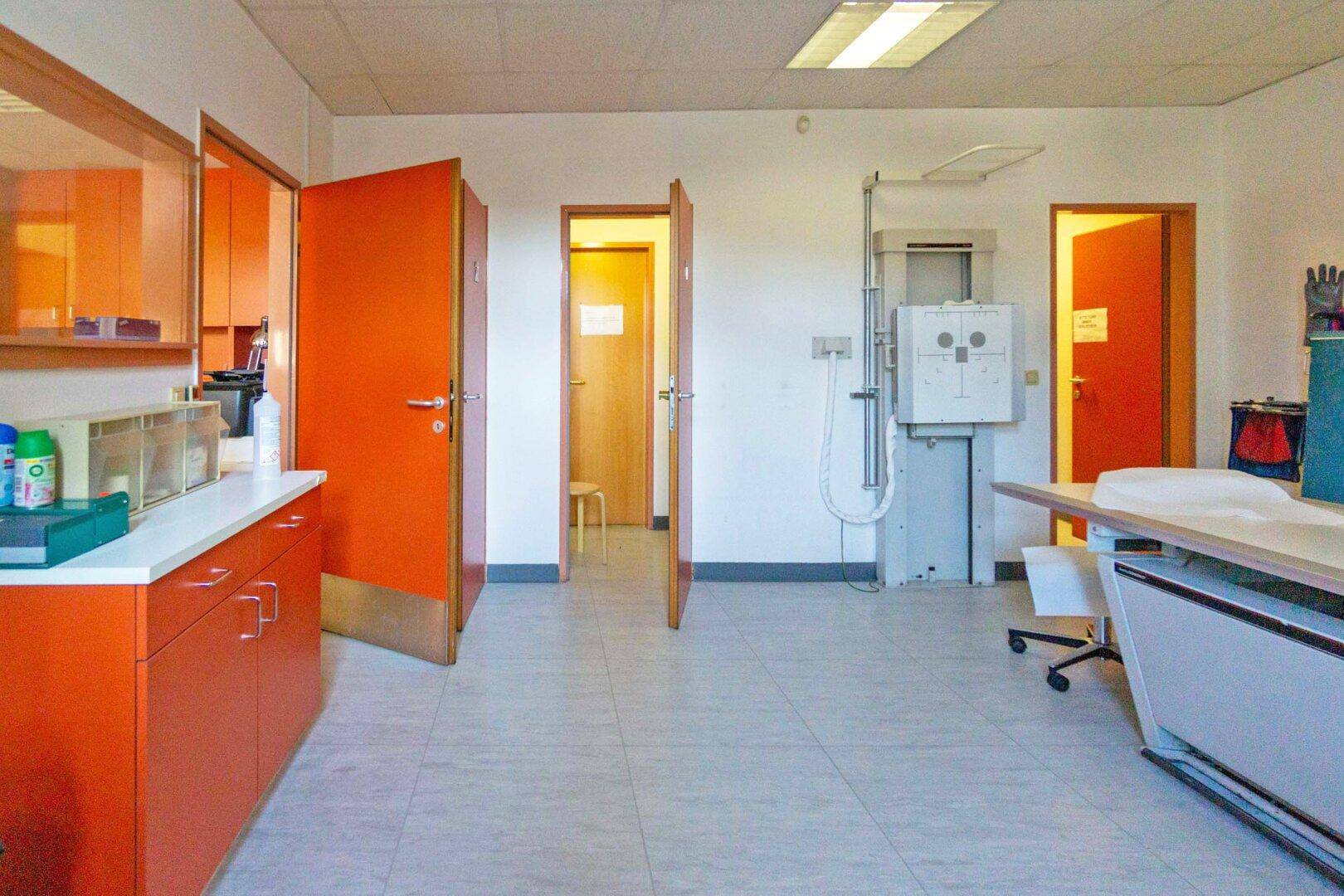 Untersuchungsraum mit medizinischen Geräten, orangefarbenen Schränken und mehreren Türen zu weiteren Räumen.