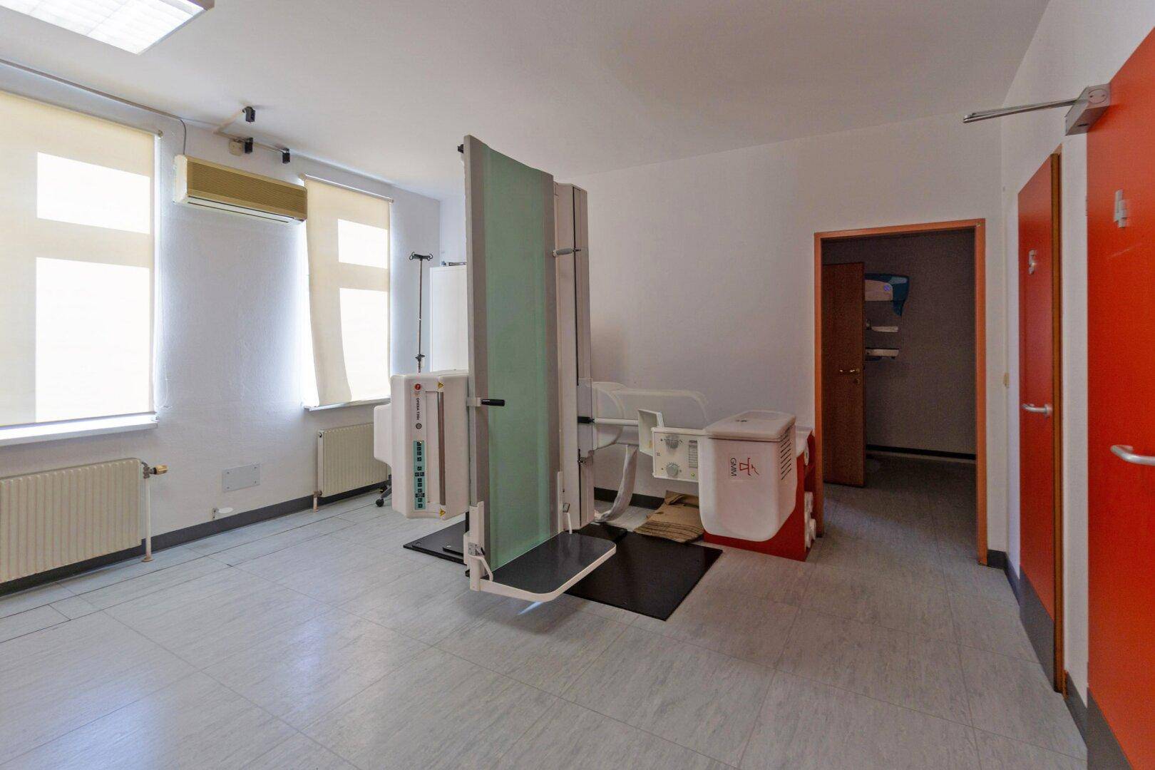 Untersuchungsraum mit spezialisierten medizinischen Geräten, Fliesenboden und Fenstern mit Rollos.