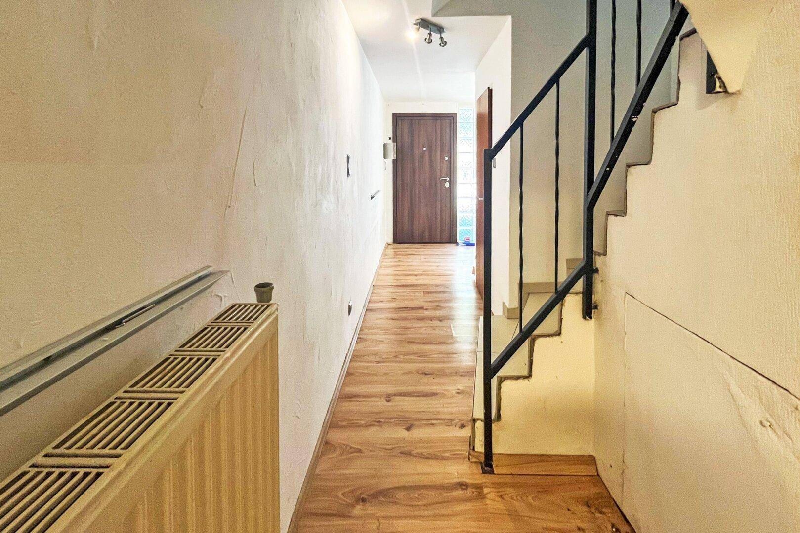 Heller Flur mit Laminatboden, Heizkörper und Treppe mit schwarzem Metallgeländer zum Eingangsbereich.