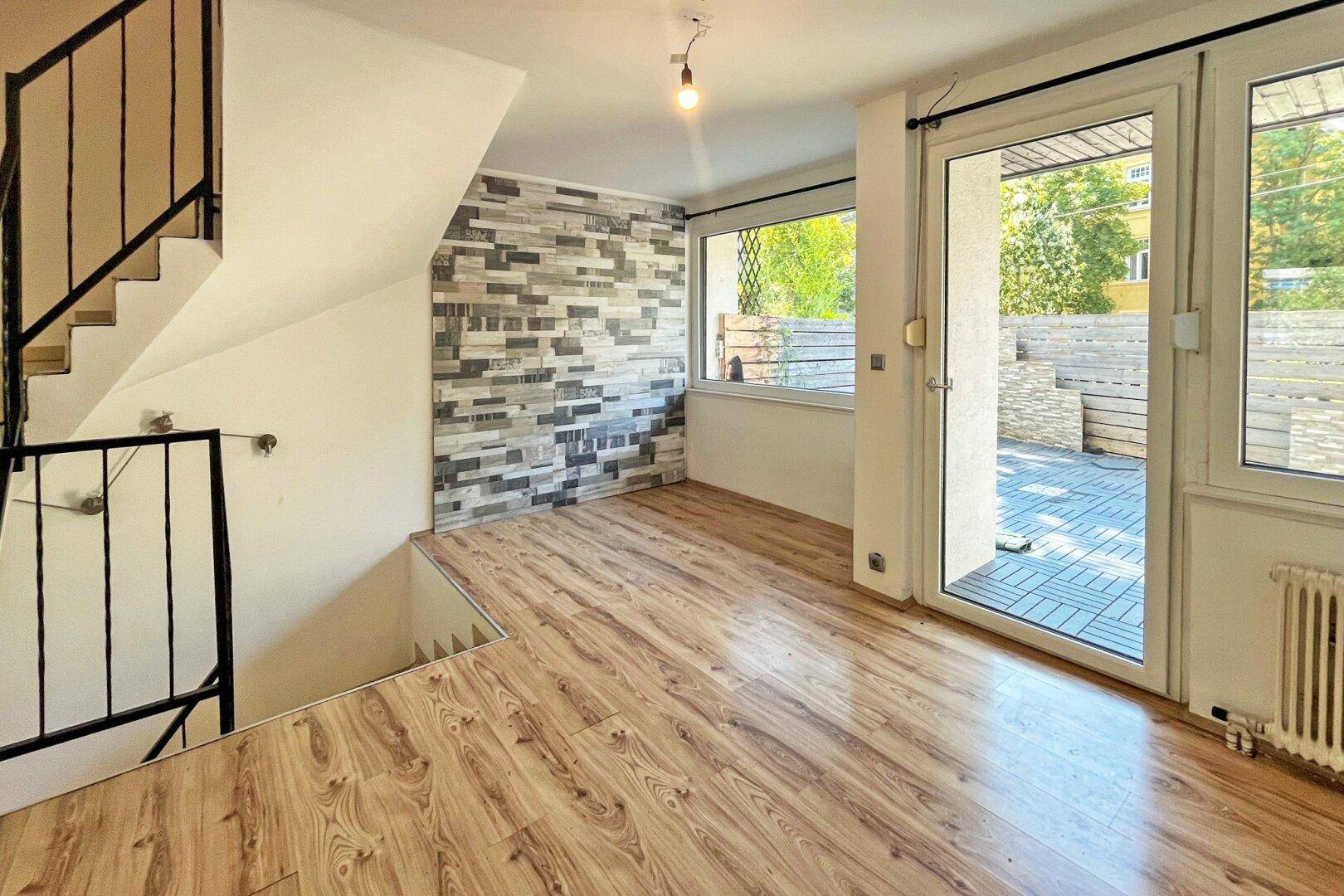 Heller Wohnbereich mit Laminatboden, dekorativer Wand und Zugang zu einer Außenterrasse.