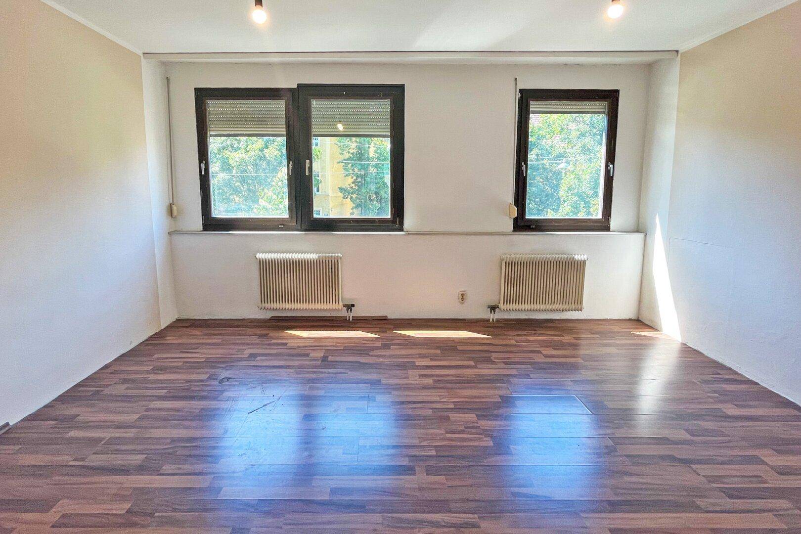 Helles Zimmer mit dunklem Laminatboden, weißen Wänden und zwei Fenstern mit Rollläden.