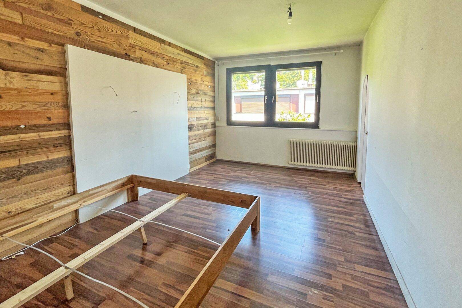 Schlafzimmer mit dunklem Laminatboden, einer Holzwand und einem Fenster mit Heizkörper.