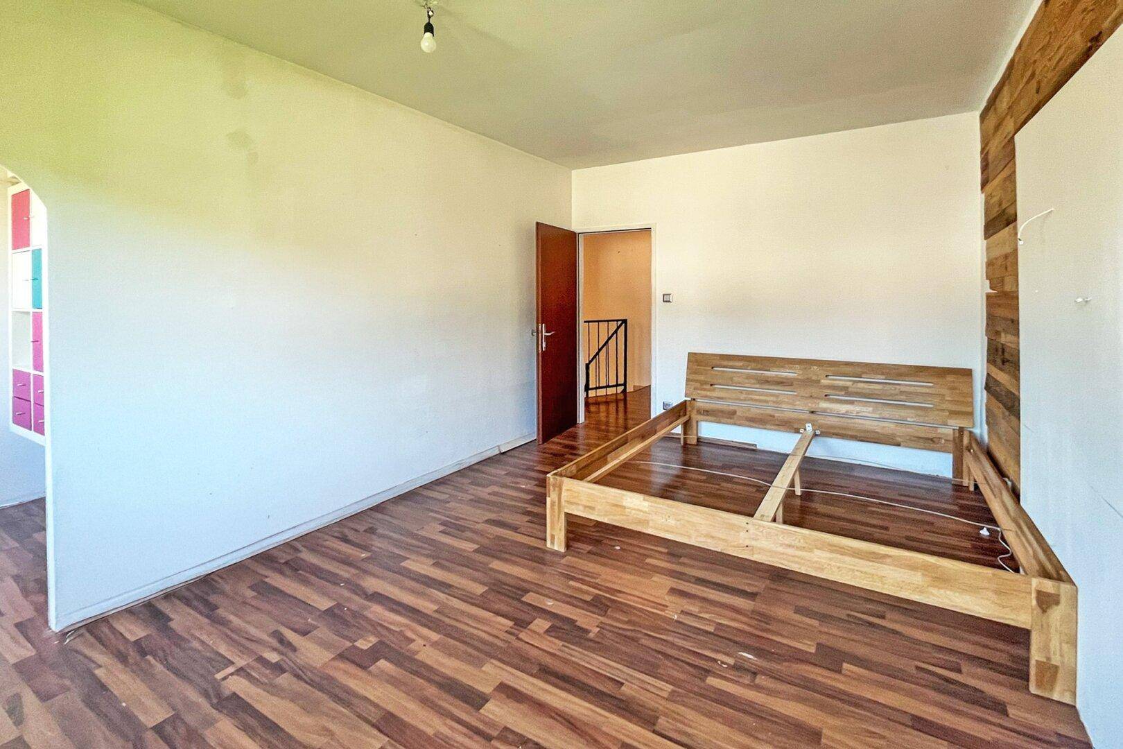 Schlafzimmer mit dunklem Laminatboden und einem Holzbettrahmen, mit Zugang zu einem weiteren Bereich.