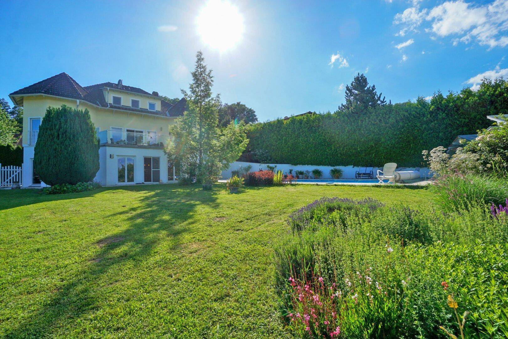 Weitläufiger, sonniger Garten mit blühenden Pflanzen und Blick auf das Haus mit Poolbereich.