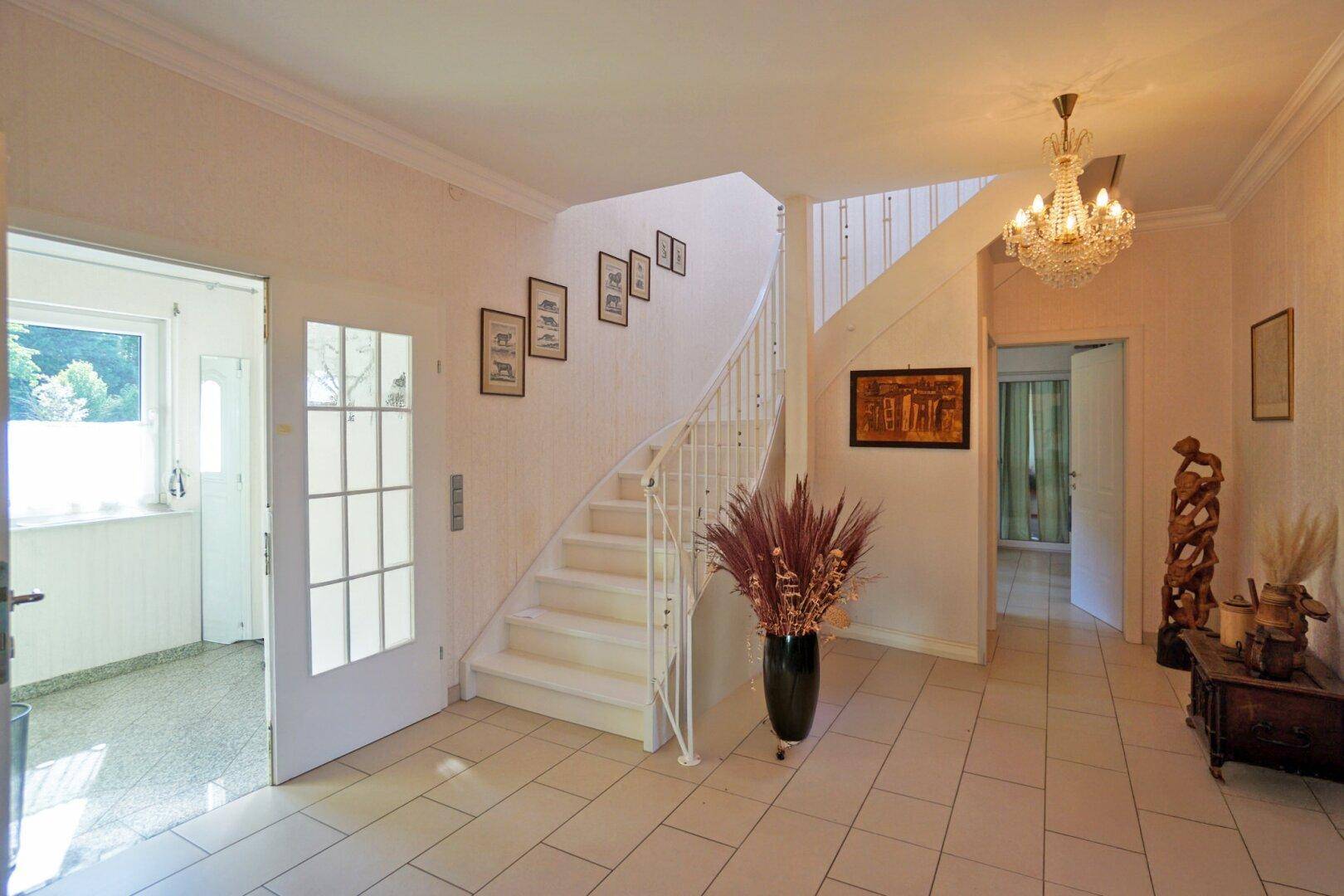 Heller Eingangsbereich mit eleganter Treppe, Fliesenboden und klassischem Kronleuchter.