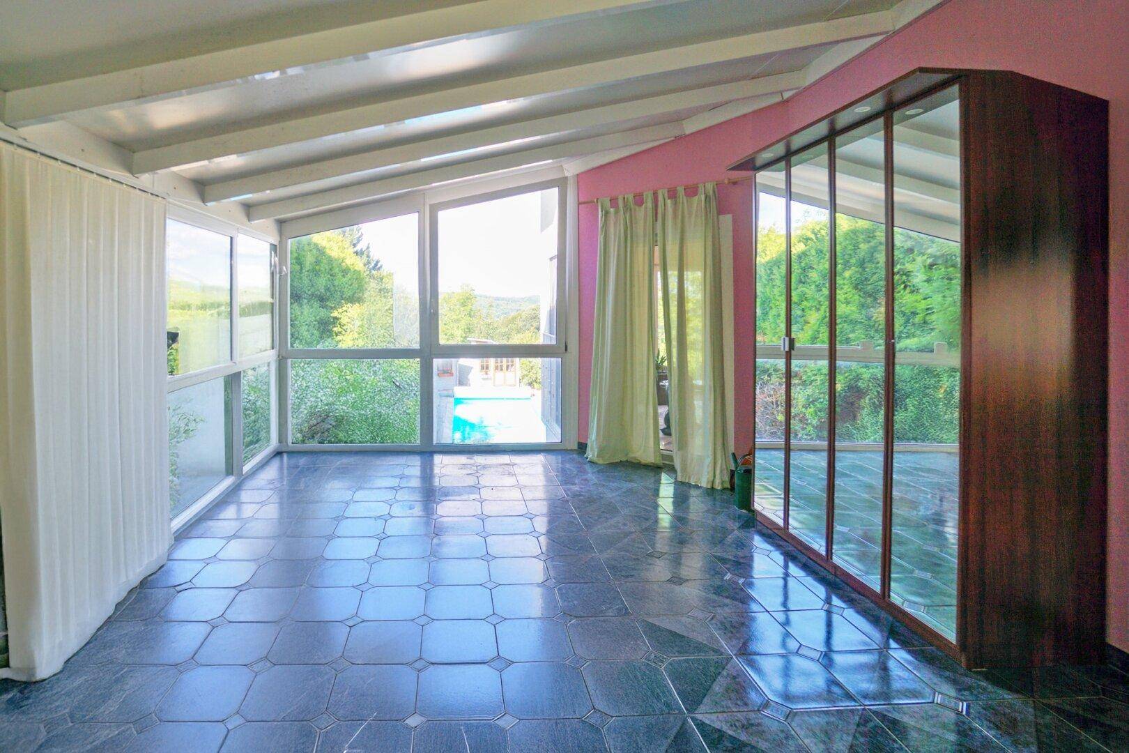 Heller Wintergarten mit großen Glasflächen, Fliesenboden und direktem Blick auf den Pool.