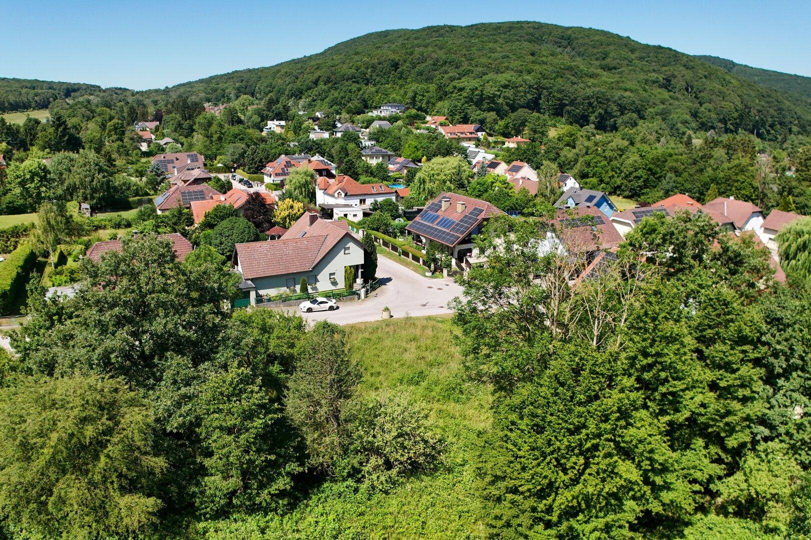 Luftaufnahme einer idyllischen Wohnsiedlung mit vielen Bäumen und grünen Flächen, umgeben von bewaldeten Hügeln.