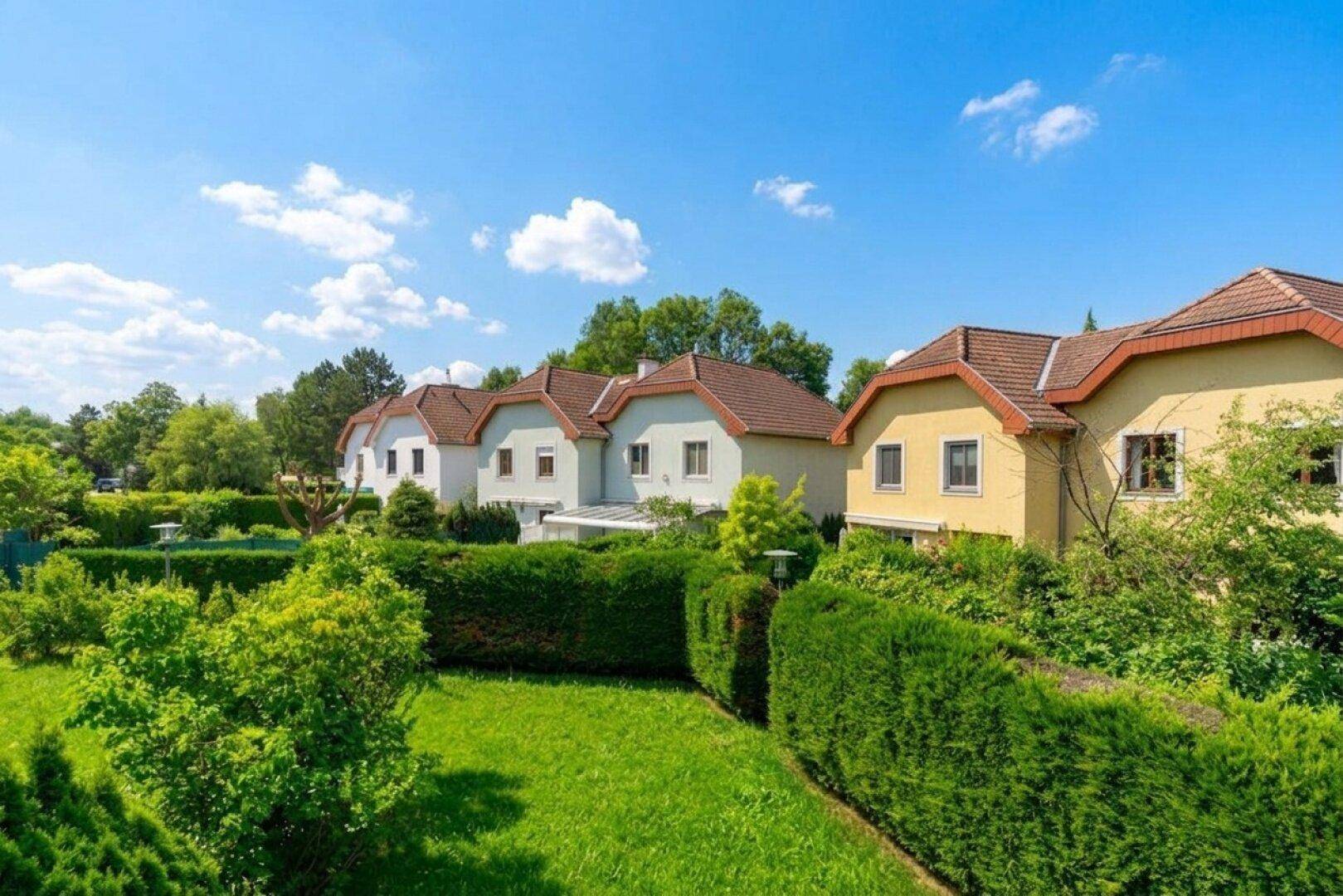 Reihenhäuser mit gepflegten Gärten unter blauem Himmel, eingebettet in eine grüne Umgebung.