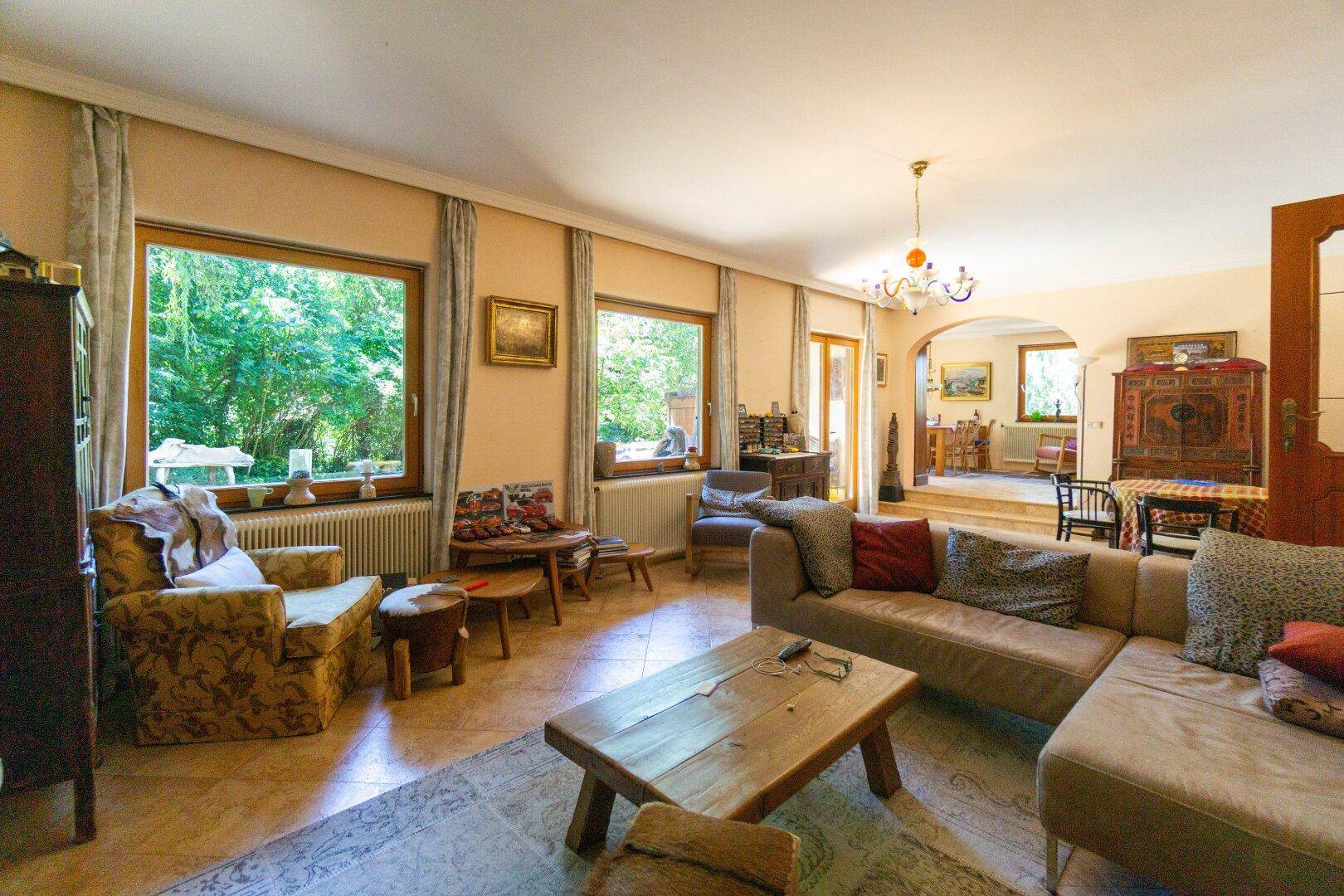 Geräumiges Wohnzimmer mit beigem Sofa, Fliesenboden und großen Fenstern mit Blick ins Grüne.