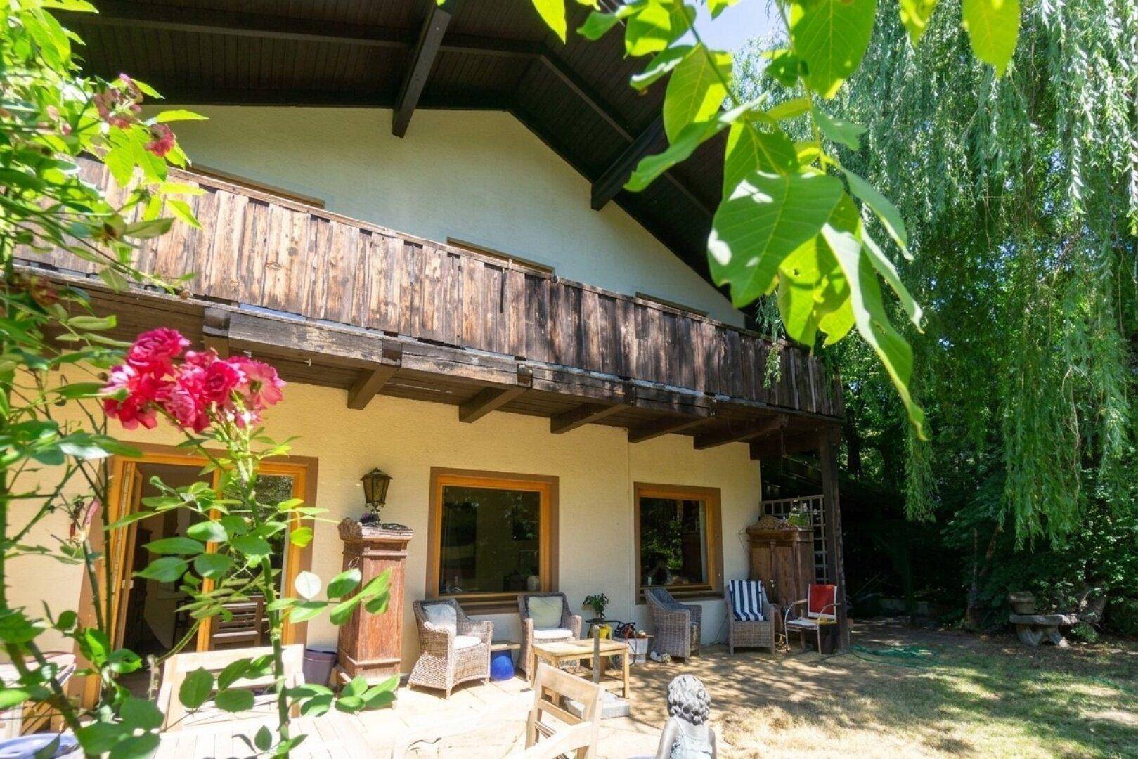 Gemütliche Terrasse mit Sitzgelegenheiten und darüberliegendem Holzbalkon, umgeben von blühenden Pflanzen.