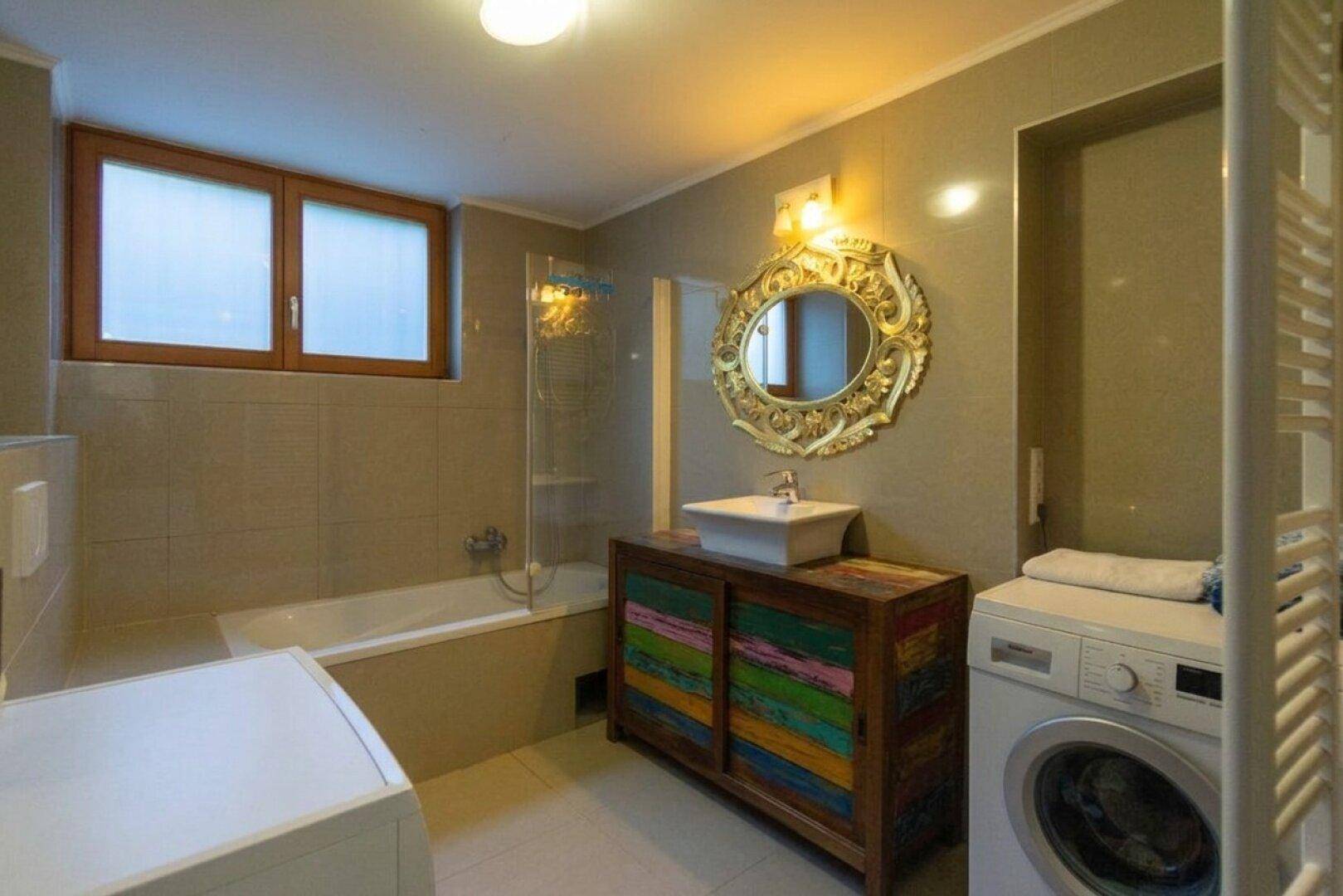 Modernes Badezimmer mit grauen Fliesen, Badewanne, Waschbecken und Waschmaschine, inklusive Fenster.