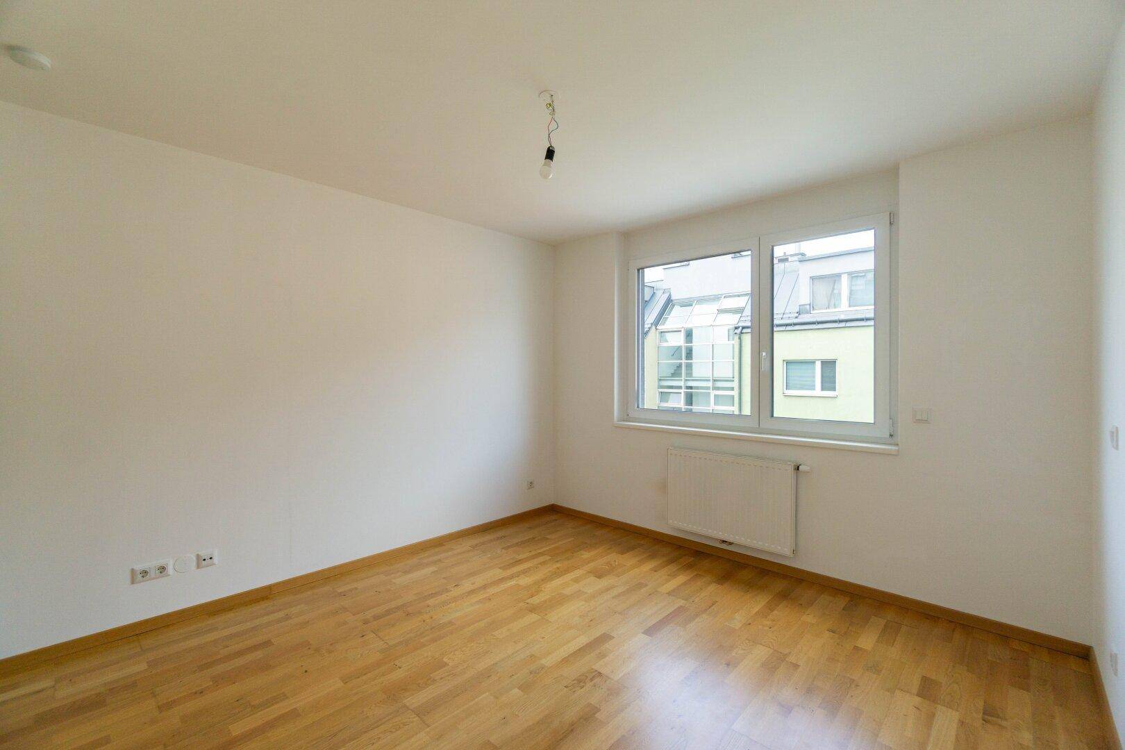 Leeres Schlafzimmer mit hellem Parkettboden und großem Fenster, das viel Tageslicht hereinlässt.