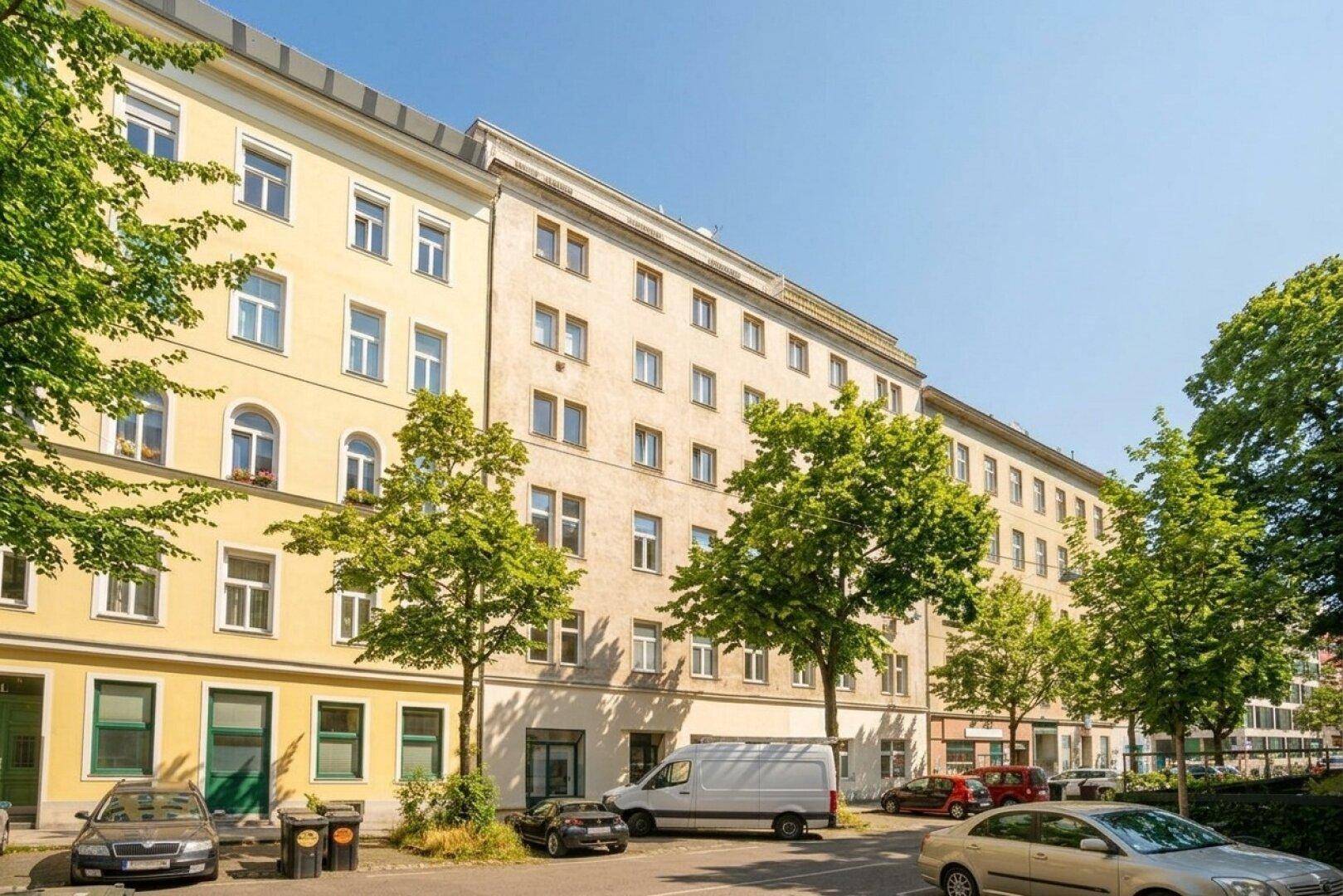 Ansicht der Wohngebäude entlang der Straße mit parkenden Autos und grünen Bäumen unter blauem Himmel.