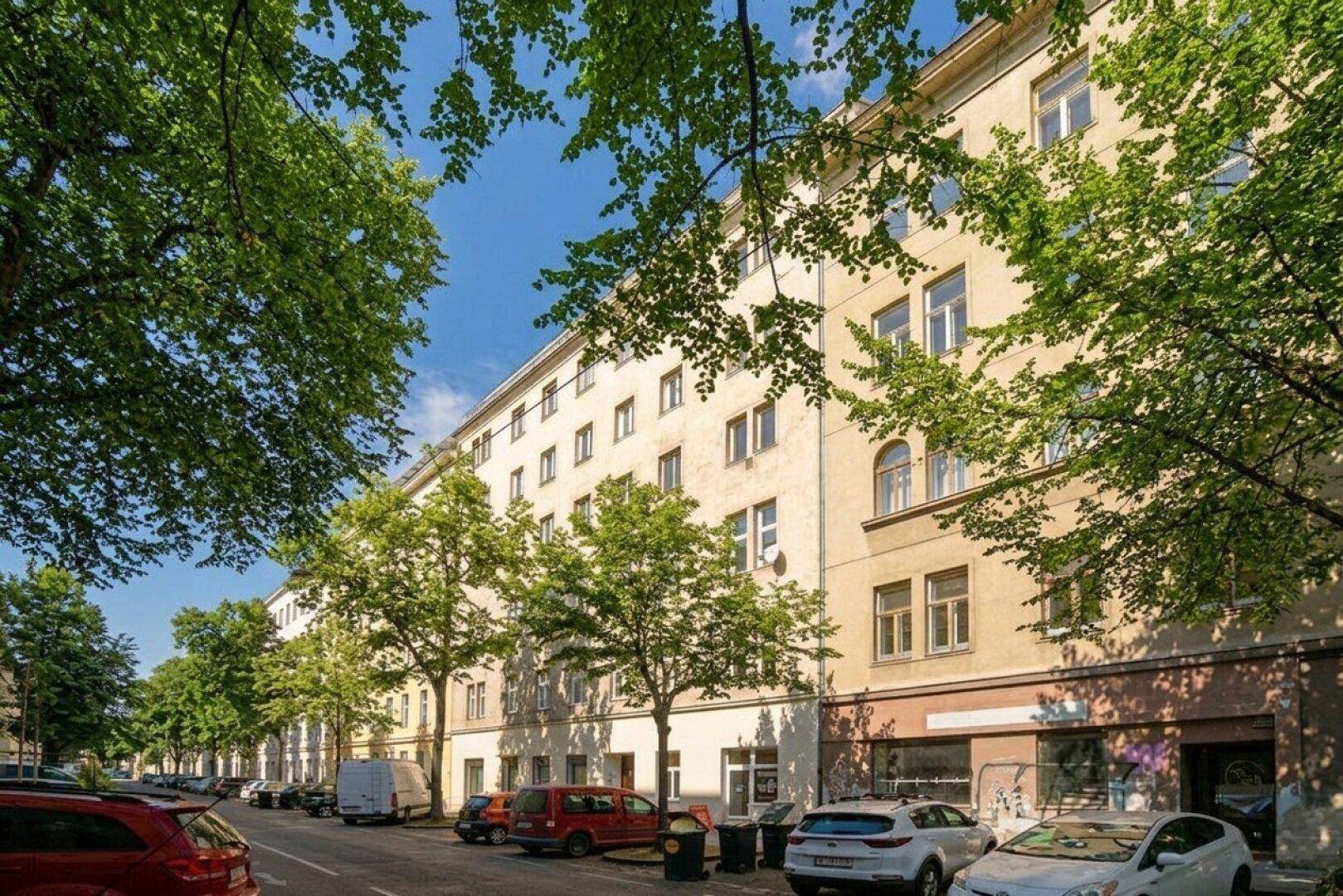 Lange Reihe von Wohngebäuden an einer städtischen Straße, gesäumt von Bäumen und parkenden Fahrzeugen.