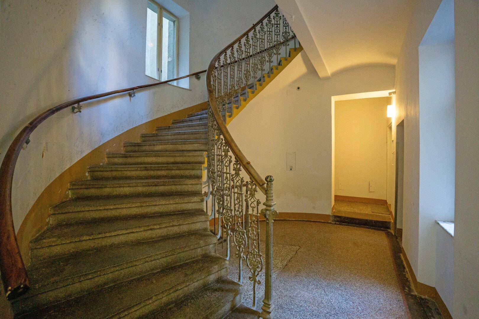 Historisches Treppenhaus mit geschwungenem Geländer, Steinstufen und einem Fenster für natürliches Licht.