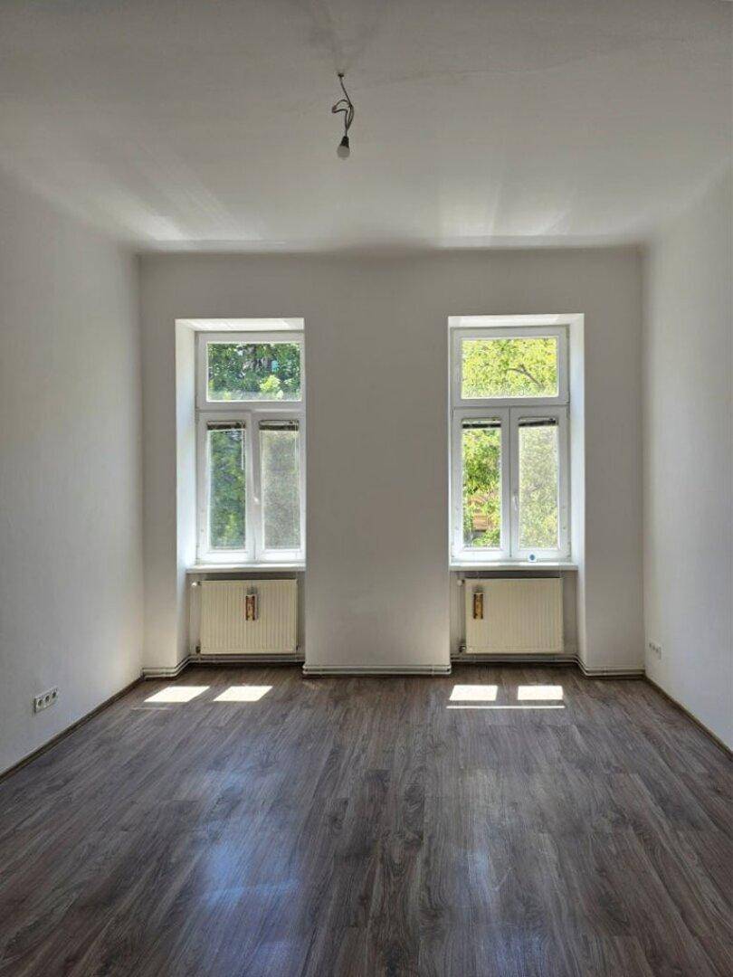 Leeres Zimmer mit dunklem Laminatboden, weißen Wänden und zwei Fenstern mit Blick ins Grüne.