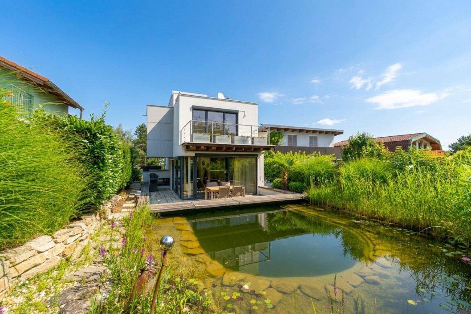 Modernes Haus mit großzügiger Terrasse am Teich, umgeben von üppigem Grün.