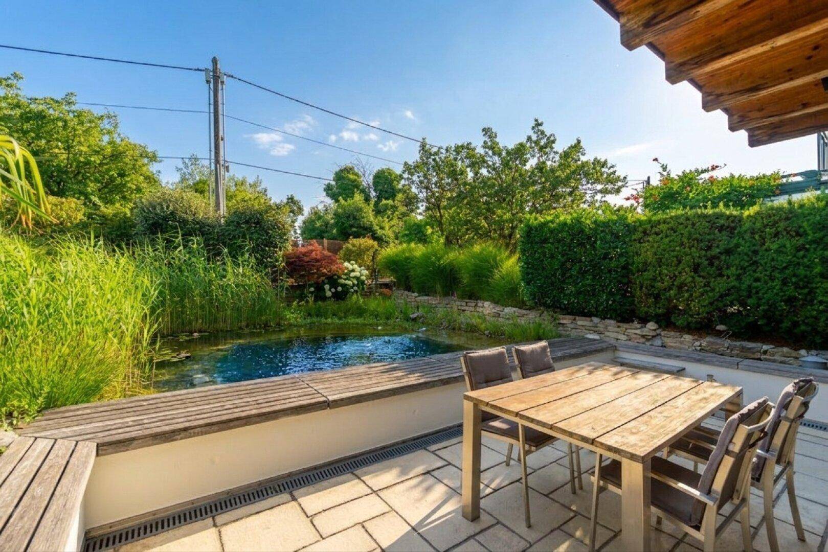 Sonnige Terrasse mit Esstisch, Holzbank und Blick auf den naturnahen Teich im Garten.
