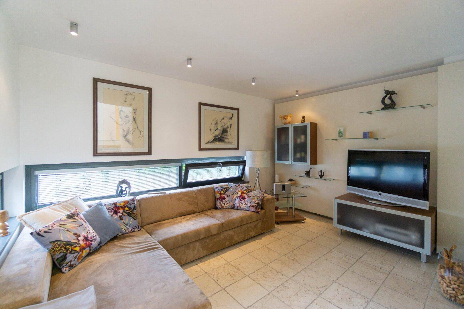 Gemütliches Wohnzimmer mit großem Sofa, Fliesenboden und niedrigen Fenstern für viel Licht.