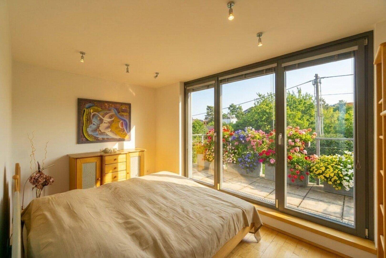 Helles Schlafzimmer mit Holzboden und großem Balkonzugang, geschmückt mit bunten Blumen.
