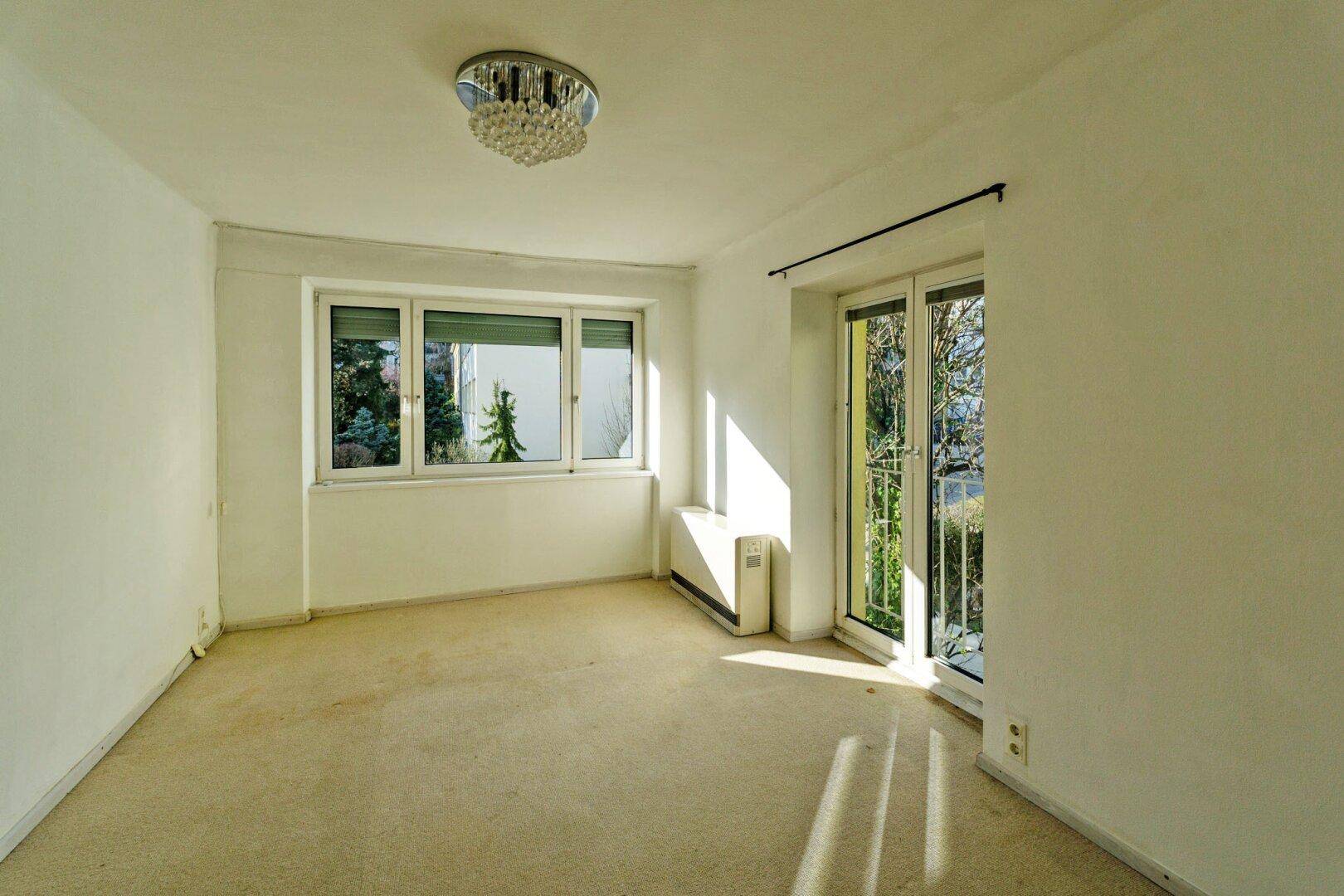 Heller Raum mit Teppichboden, großen Fenstern, Heizkörper und Zugang zu einem französischen Balkon mit Grünblick.