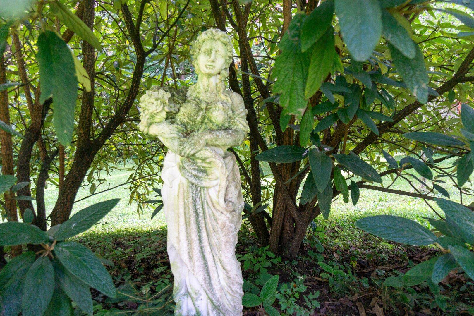 Verwitterte Steinstatue einer Frau mit Blumen, umgeben von dichtem Grün und Bäumen in einem idyllischen Garten.