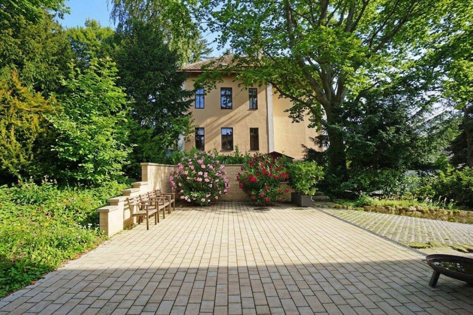 Geräumiger gepflasterter Vorplatz mit Sitzbänken, umgeben von üppigem Grün und hohen Bäumen.